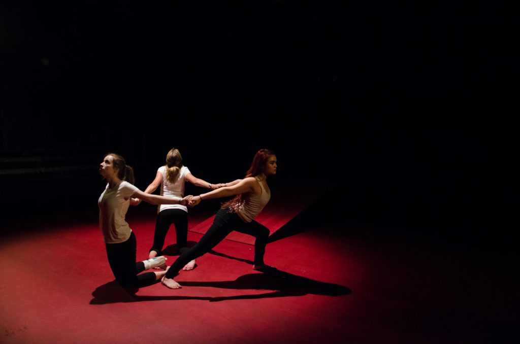 3 young actors with arms locked pull away from each other. The ground is painted red. There is a spotlight on the actors, the rest is pitch black.
